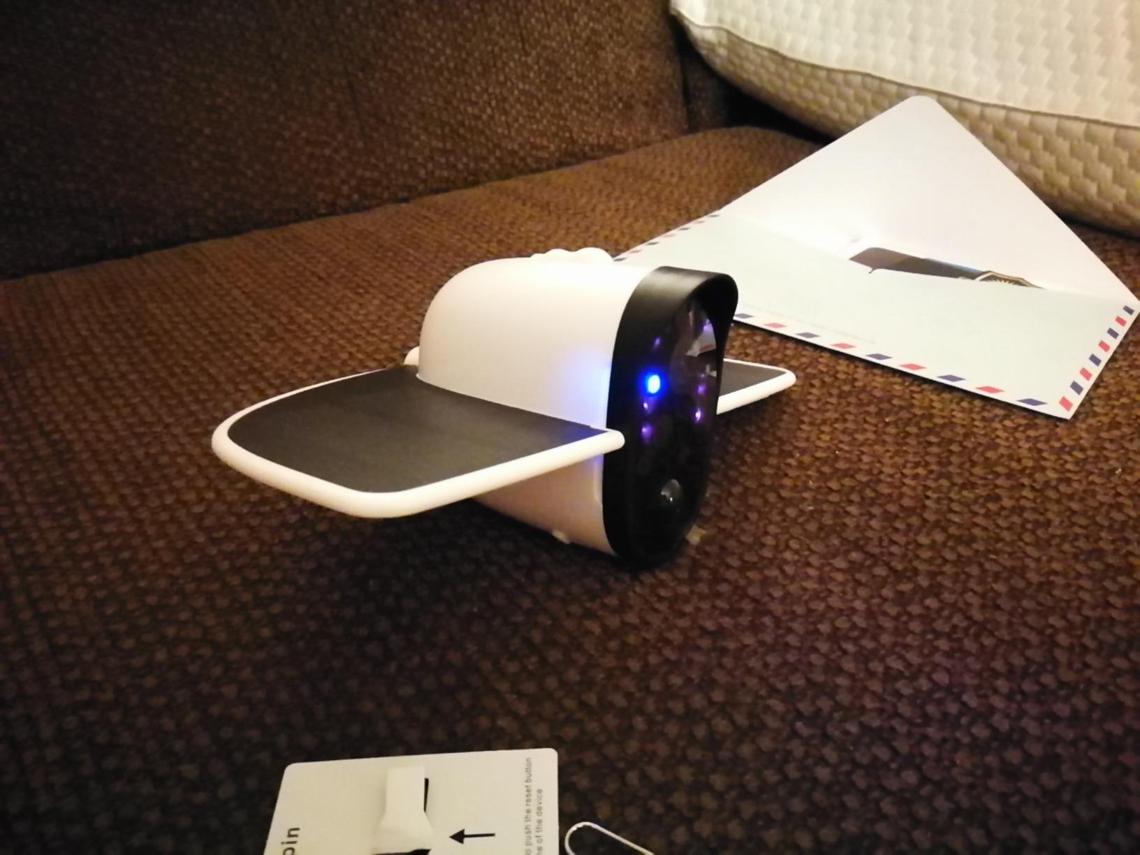 White smart home security camera with wings on a brown textured couch next to papers and a card