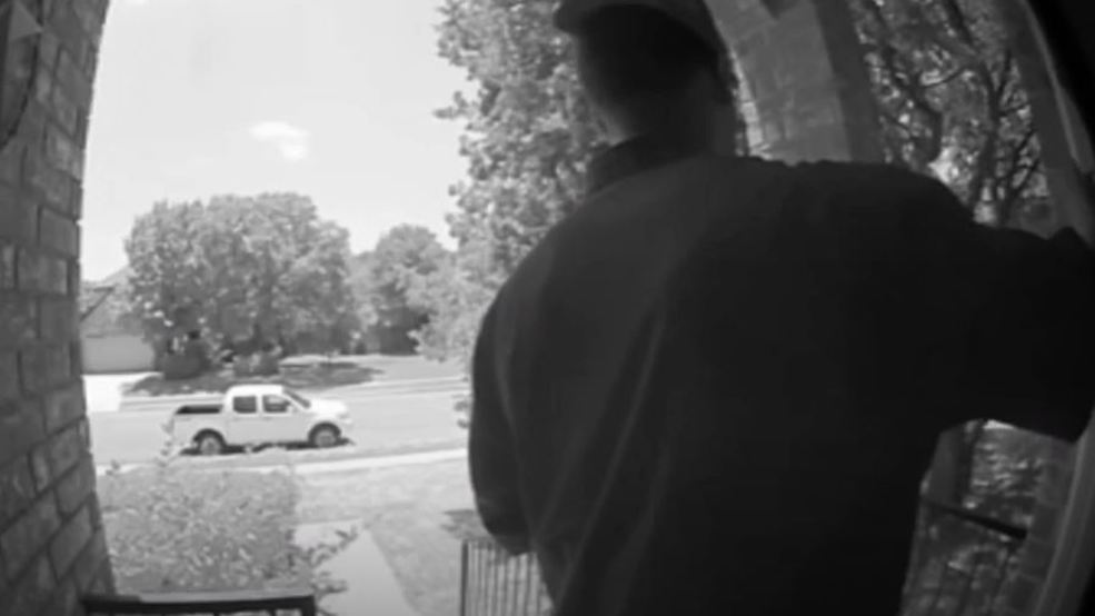 Black and white security camera image of a man standing on a front porch near an open door with a white pickup truck on the street