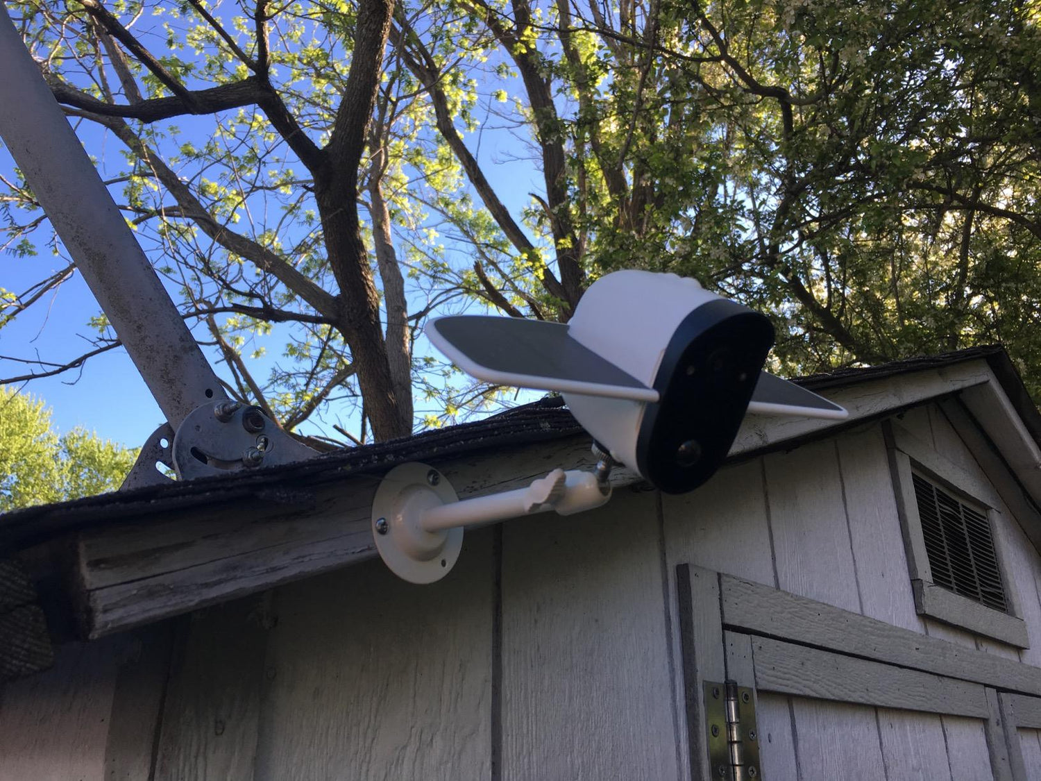White security camera mounted on the corner of a wooden shed roof with green trees in background