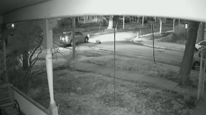 Nighttime street scene captured by porch security camera showing a moving SUV and parked car