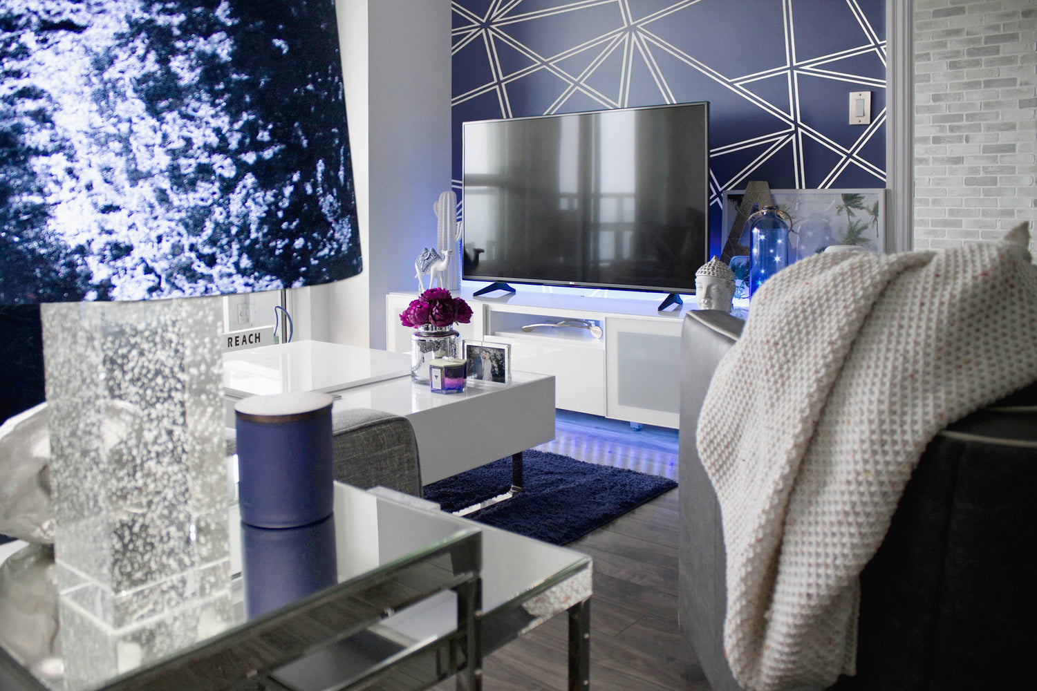 Modern living room with large flat-screen TV, geometric navy blue wallpaper, white furniture, and cozy blanket