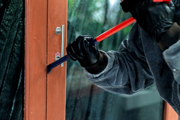Masked burglar wearing black gloves using red crowbar to pry open wooden door lock