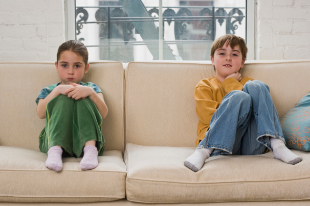 Two kids sitting separately on a beige couch in a bright living room with a window behind