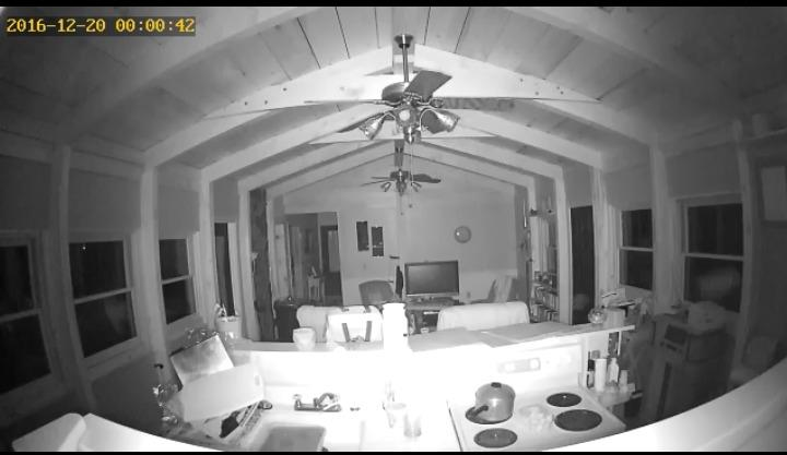 Black and white night view of a living room and kitchen with ceiling fans and TV inside a wooden cabin