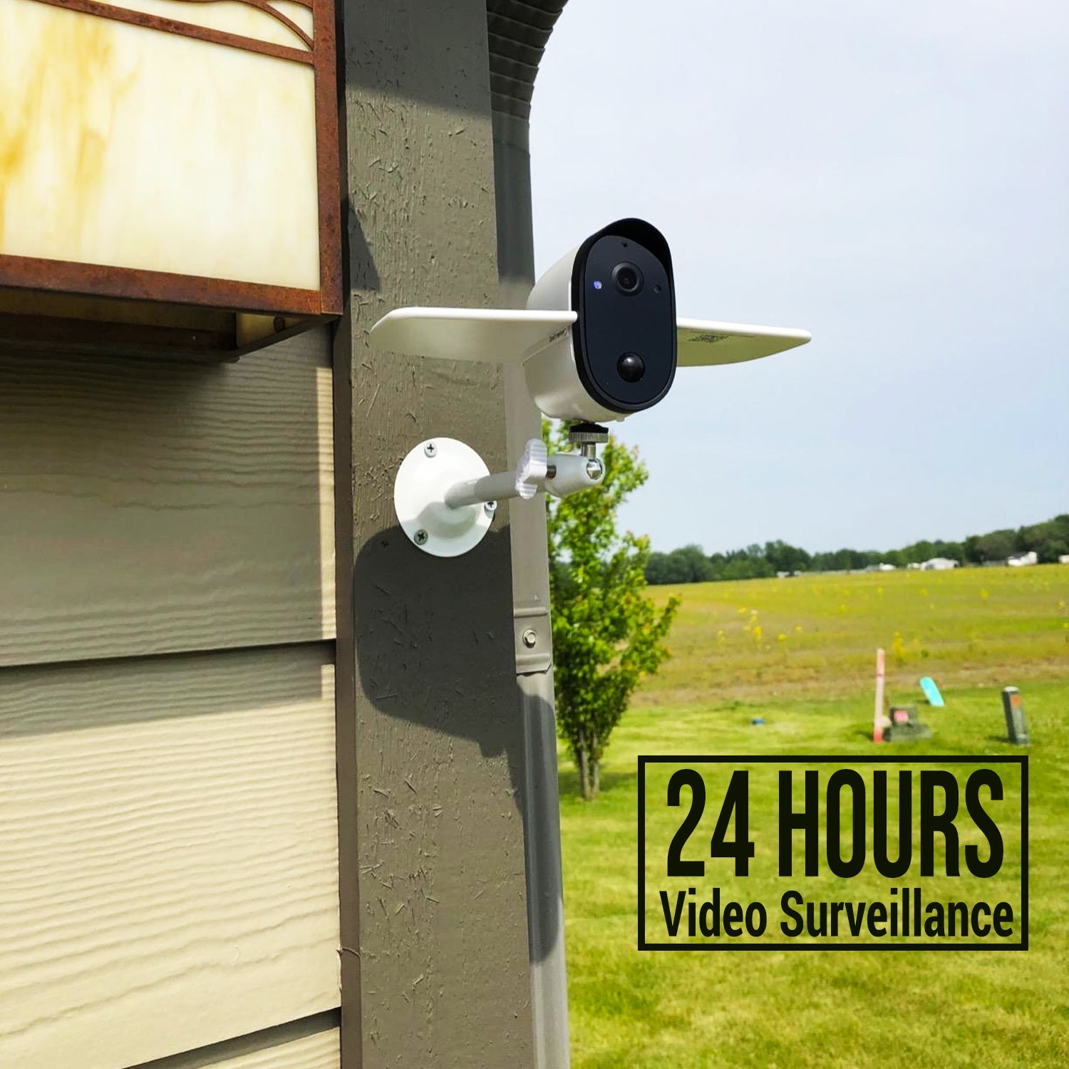 Outdoor security camera mounted on house corner with green field and 24 hours video surveillance text