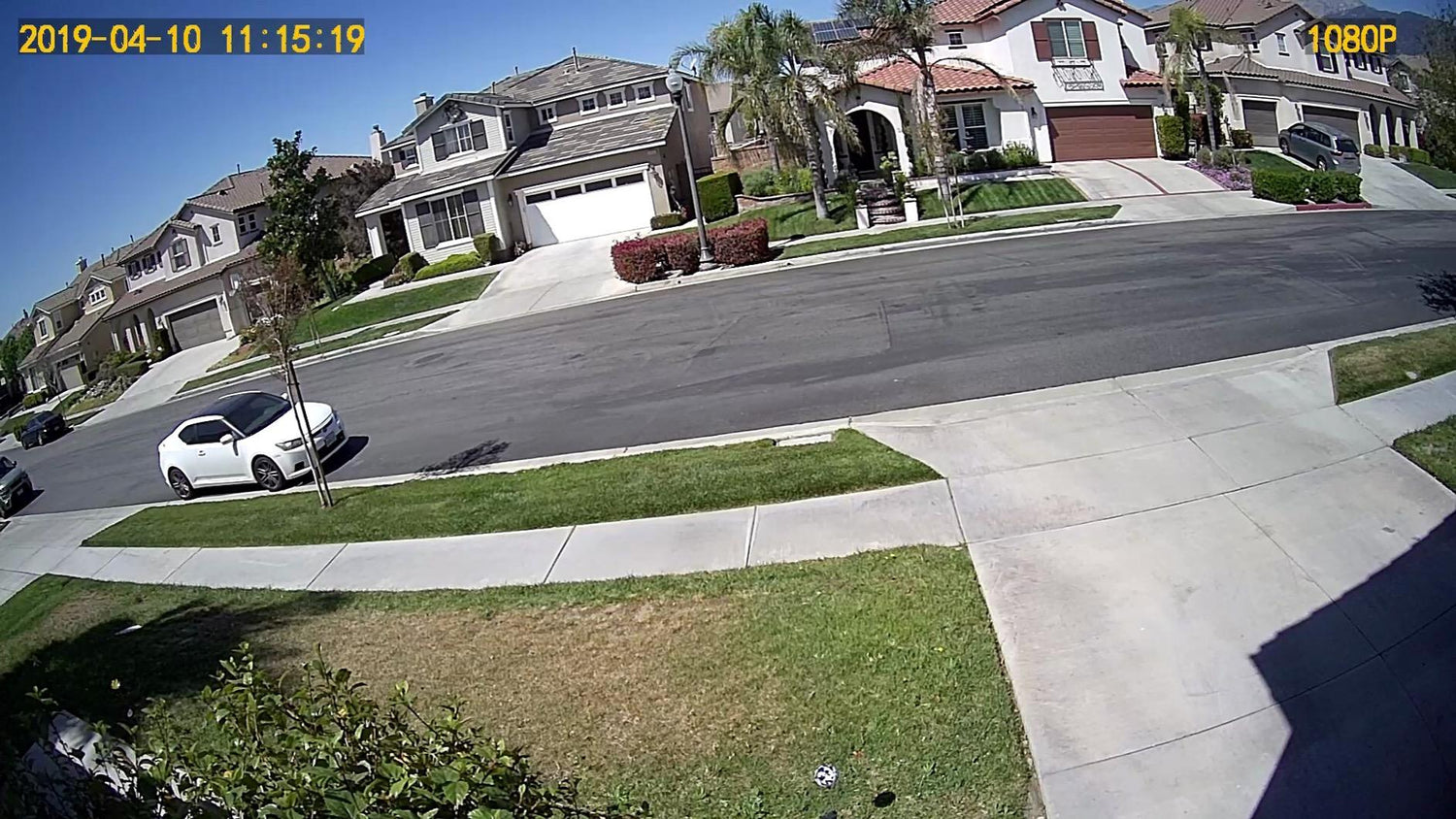 Suburban neighborhood street with modern houses, green lawns, parked white car, and clear blue sky