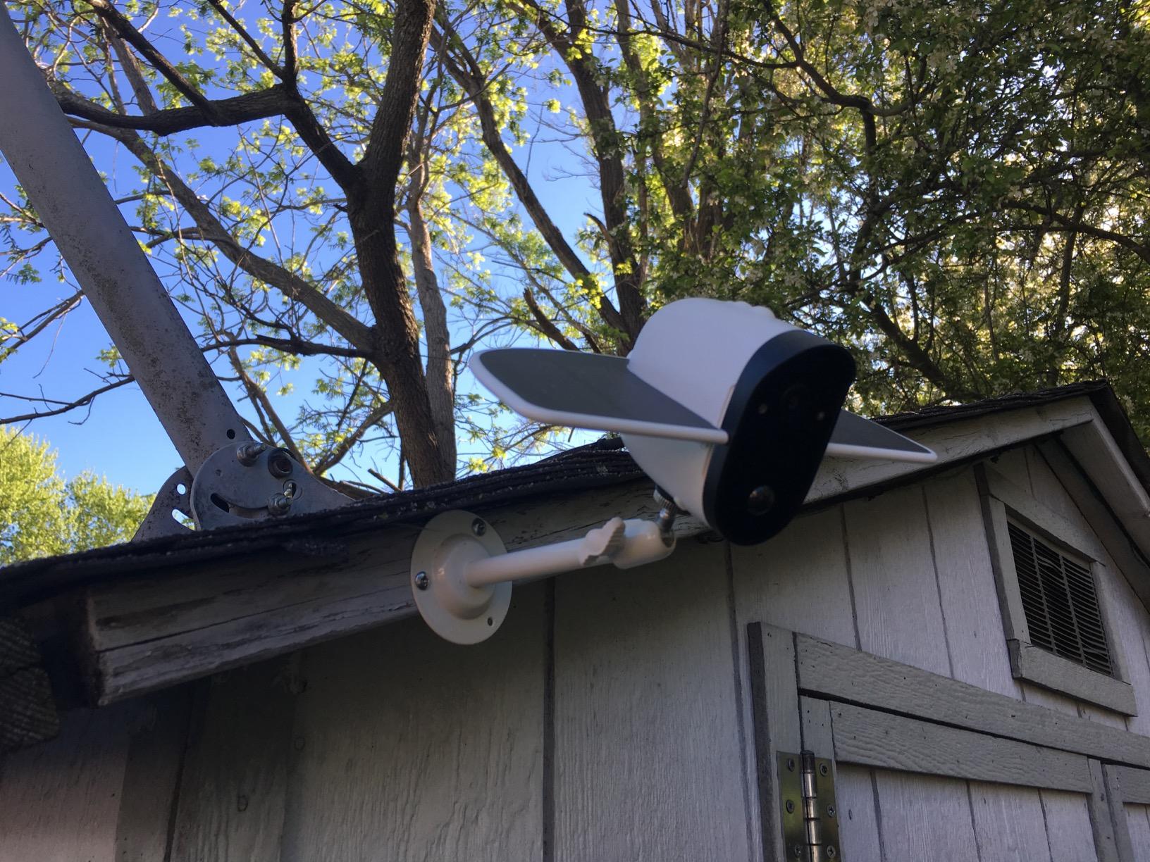 White security camera mounted on the corner of a wooden shed roof with green trees in background