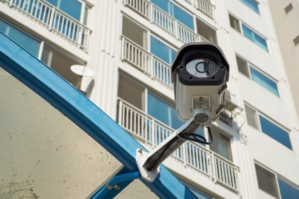 Close-up of a white security camera mounted on a blue metal structure with an apartment building in the background