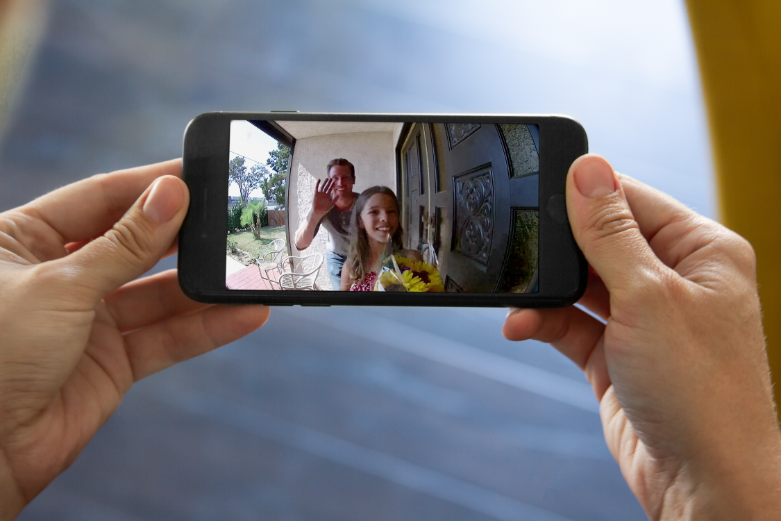 View of young man and girl with flowers on smart doorbell camera screen, hands holding smartphone