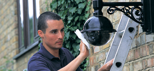 Technician installing or cleaning a black security camera mounted on a brick wall using a ladder