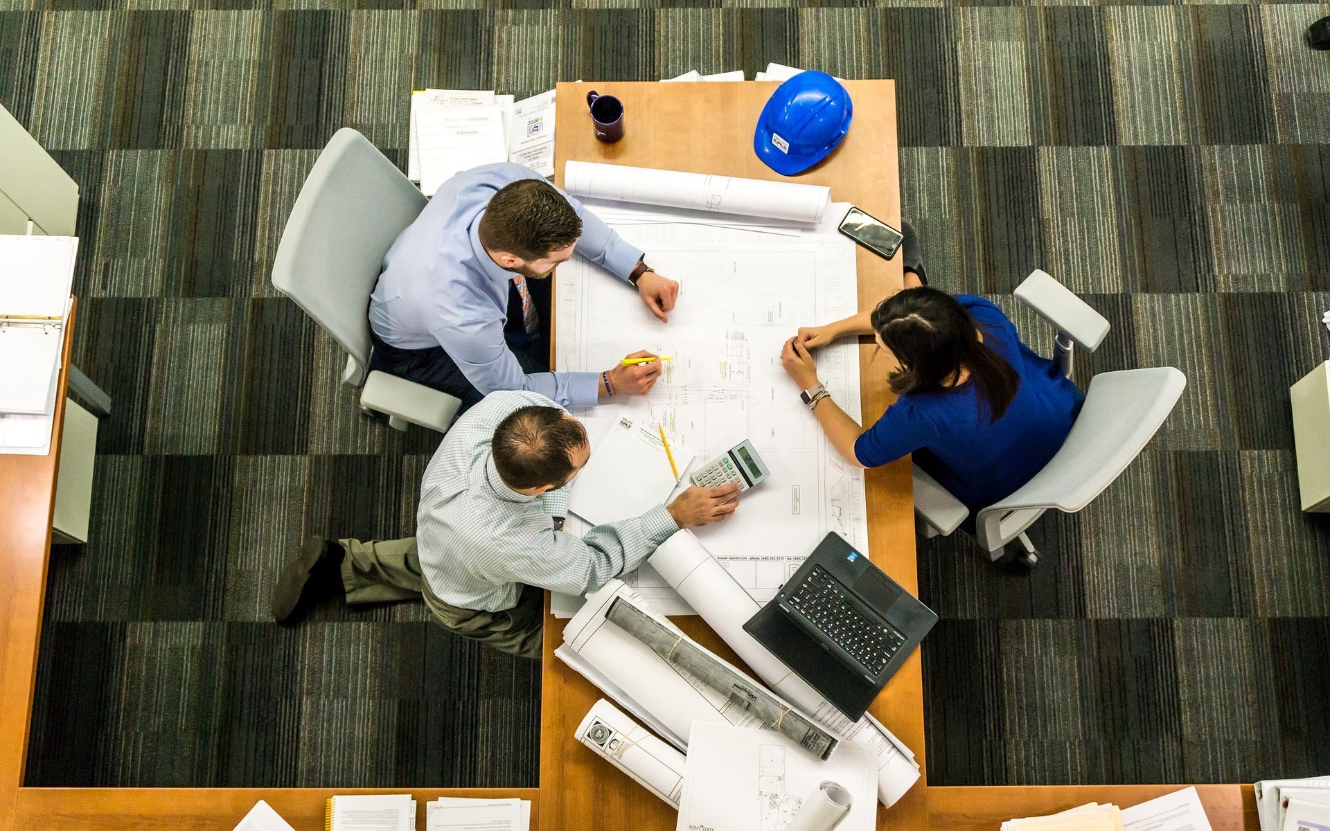 Three architects discussing blueprints at office table with laptop, calculator, and blue safety helmet
