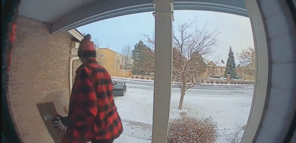 Person in red plaid jacket and hat stealing package from snow-covered porch in suburban neighborhood