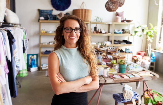 Confident young woman standing with arms crossed in a bright boutique retail store with clothing and handmade goods